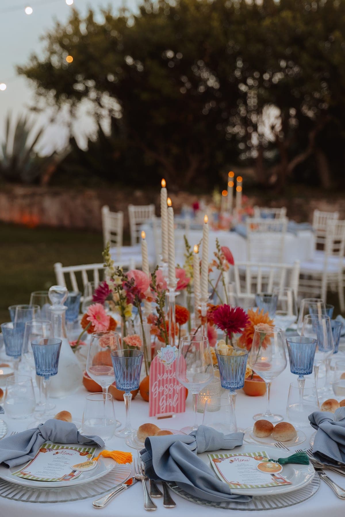 Tavola apparecchiata al tramonto con candele, fiori colorati e bicchieri azzurri
